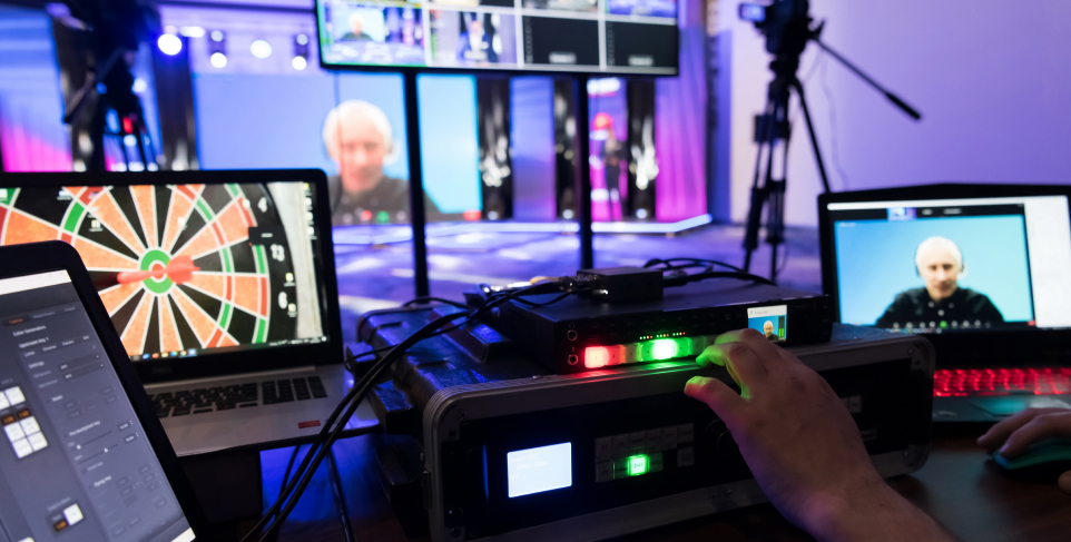 A technician's hand adjusts controls on video production equipment in a studio setting where multiple monitors and laptops display a live virtual conference with a remote speaker.