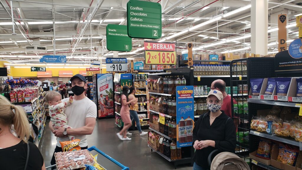 A man wearing a face mask holds a young child while standing in the snack aisle of a busy grocery store featuring Spanish-language signage and various merchandise.