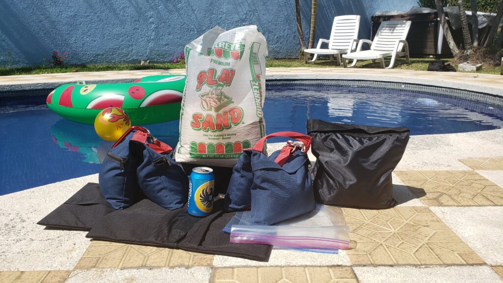 A bag of Quikrete Premium Play Sand sits by a pool alongside weighted bags, a blue soda can, and a clear plastic bag on a tiled patio.