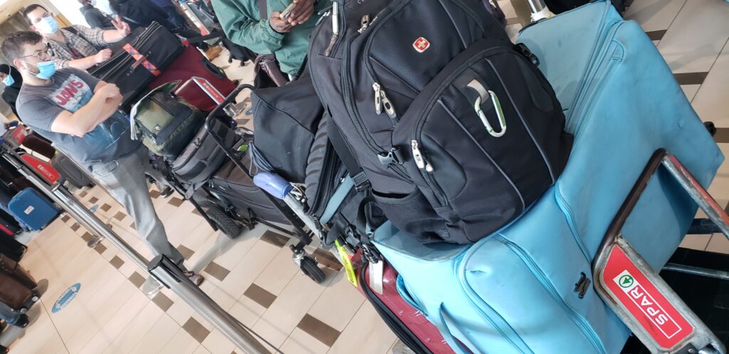 Braun video production team wait in an airport terminal with their luggage, featuring a prominent blue suitcase and black backpack stacked on a baggage cart labeled "SPAR" in the foreground.