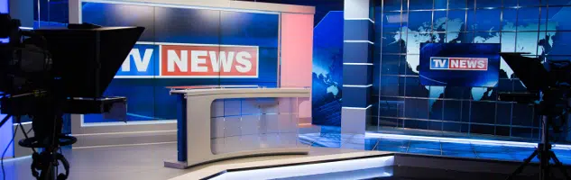 A professional television news studio features a modern anchor desk centered before large digital displays showing world maps and "TV NEWS" graphics, with broadcast cameras positioned in the foreground.