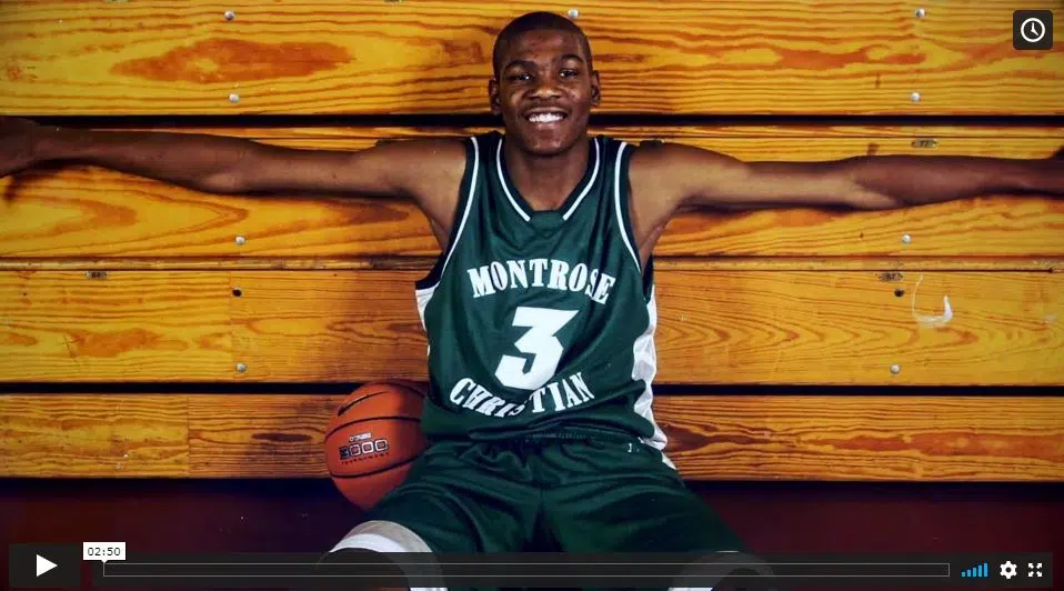 A young Kevin Durant smiles while sitting on a gym floor in his green and white Montrose Christian basketball uniform, his long arms outstretched against wooden bleachers.