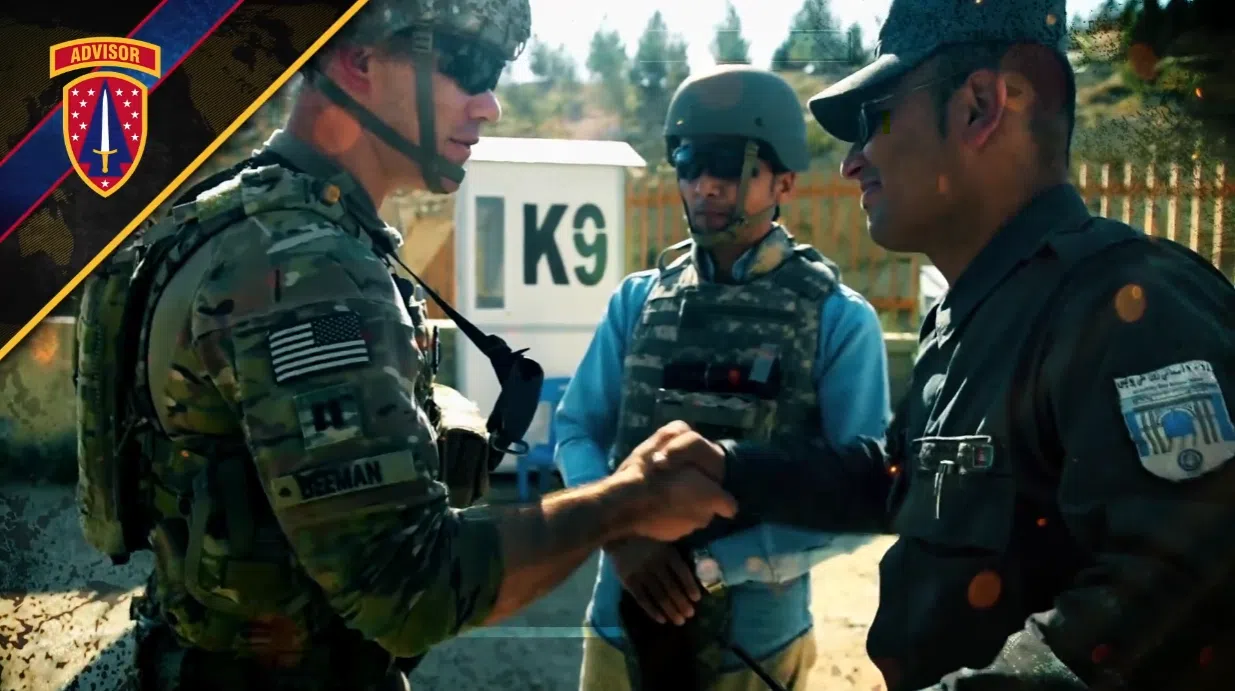 A U.S. Army Security Force Assistance Brigade (SFAB) advisor in camouflage uniform shakes hands with an Afghan partner while a third service member observes in a desert setting near a "K9" structure.