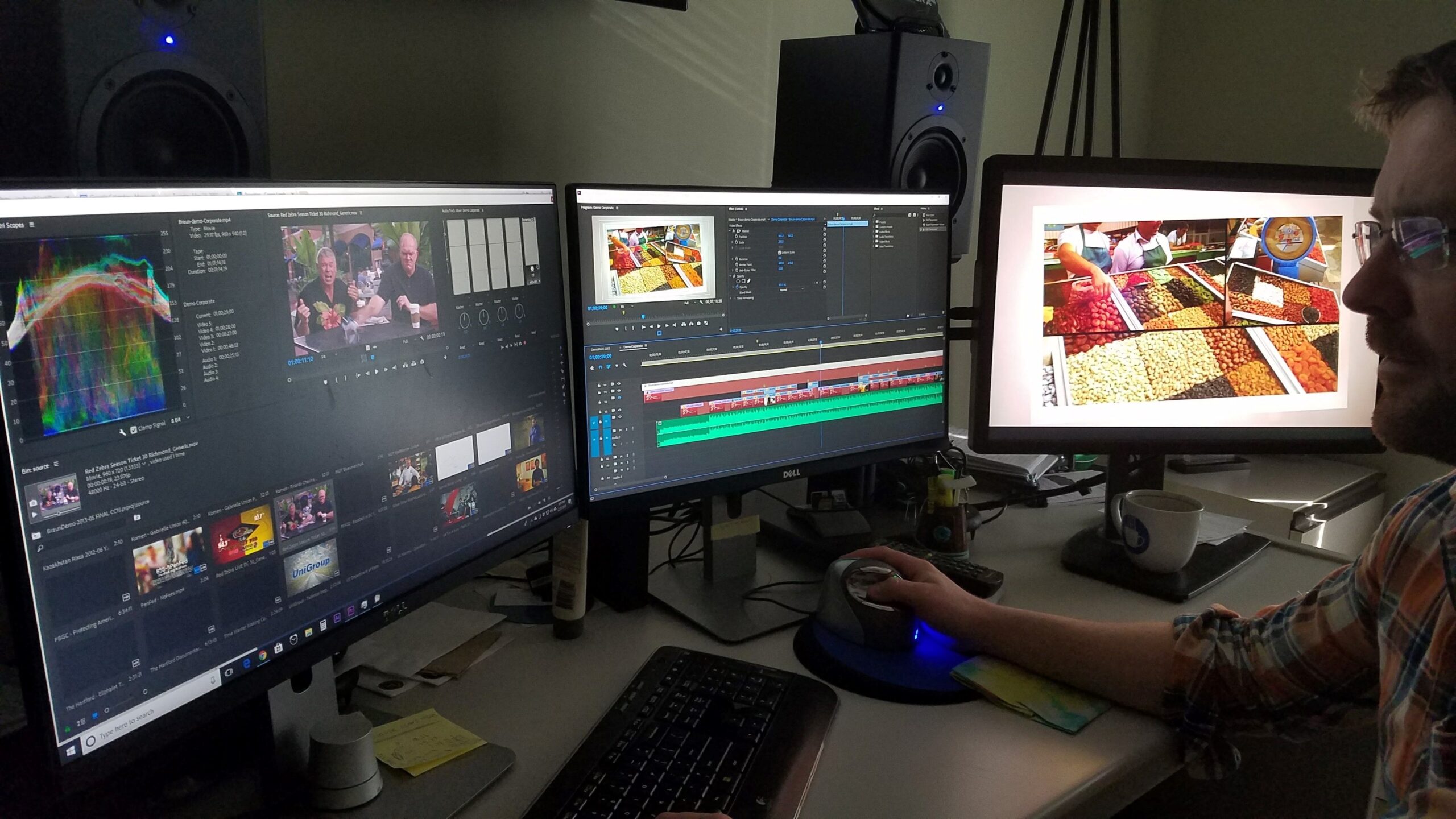 A man sits at a desk in a darkened room, editing video across three computer monitors while using a specialized trackball mouse.