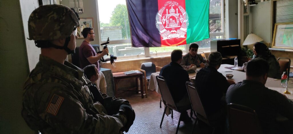 A US soldier stands in the foreground of a room where a group of people is gathered for a meeting or film production under a large flag of Afghanistan hanging in a window.
