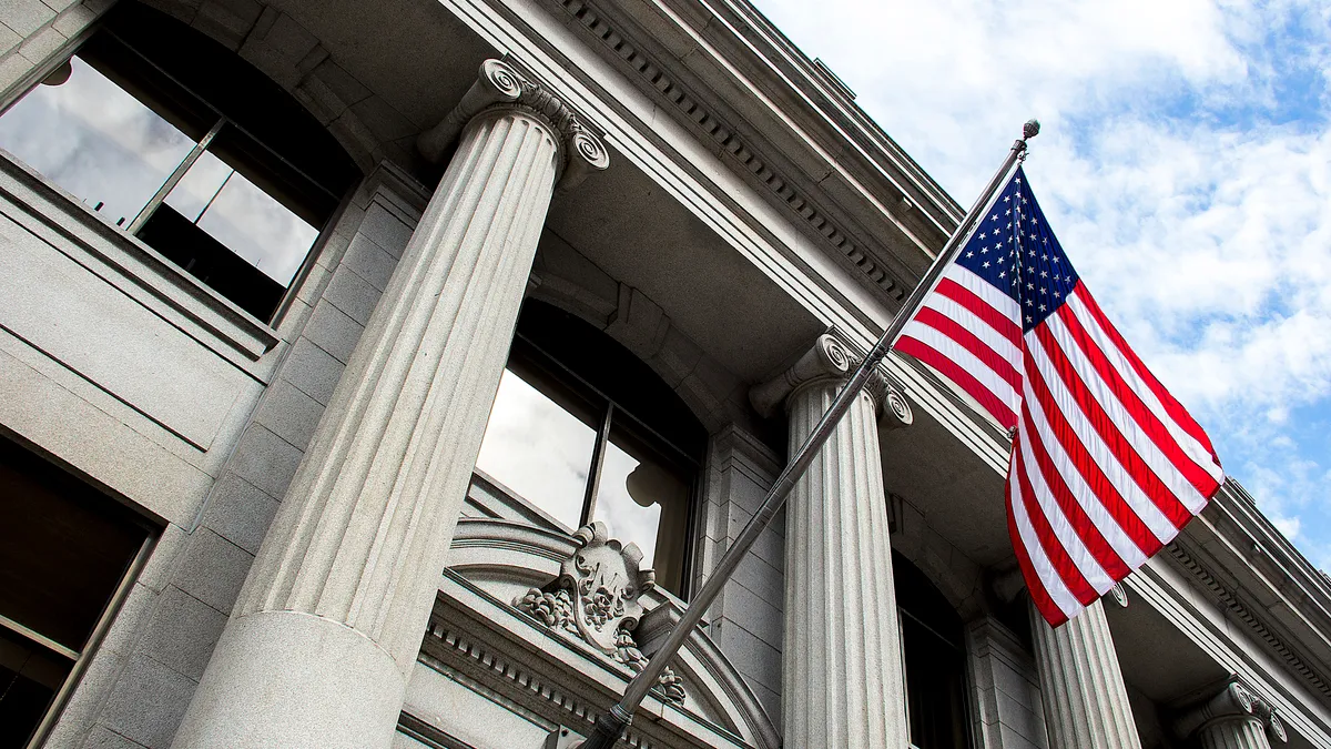 A low-angle view of a neoclassical U.S. government building featuring prominent fluted columns and an American flag waving against a blue, cloud-streaked sky.