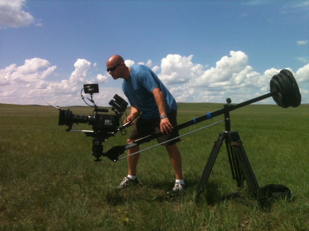Gary Braun in a blue shirt and shorts operates a professional camera mounted on a large jib crane in an open, grassy field under a bright, cloudy sky.