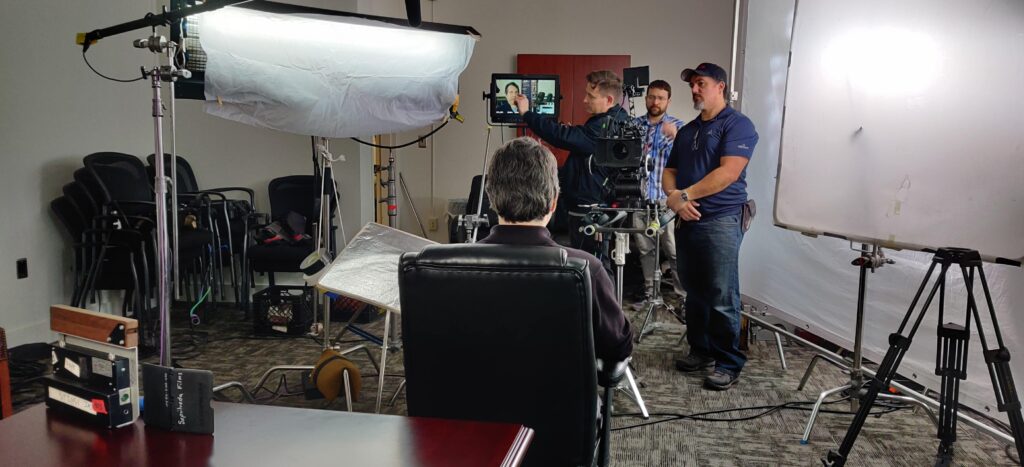 Video production crew filming an interview indoors with cameras, lighting equipment, reflectors, and a seated interview subject.