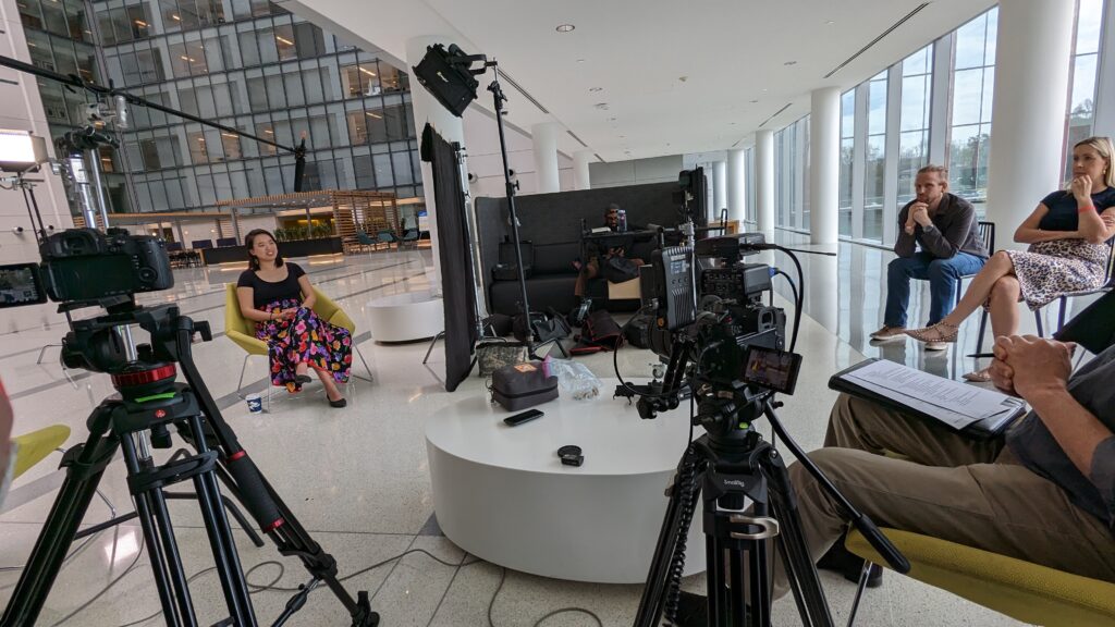 Video production crew filming a seated interview in a bright indoor lobby with cameras, lighting equipment, and crew members visible.