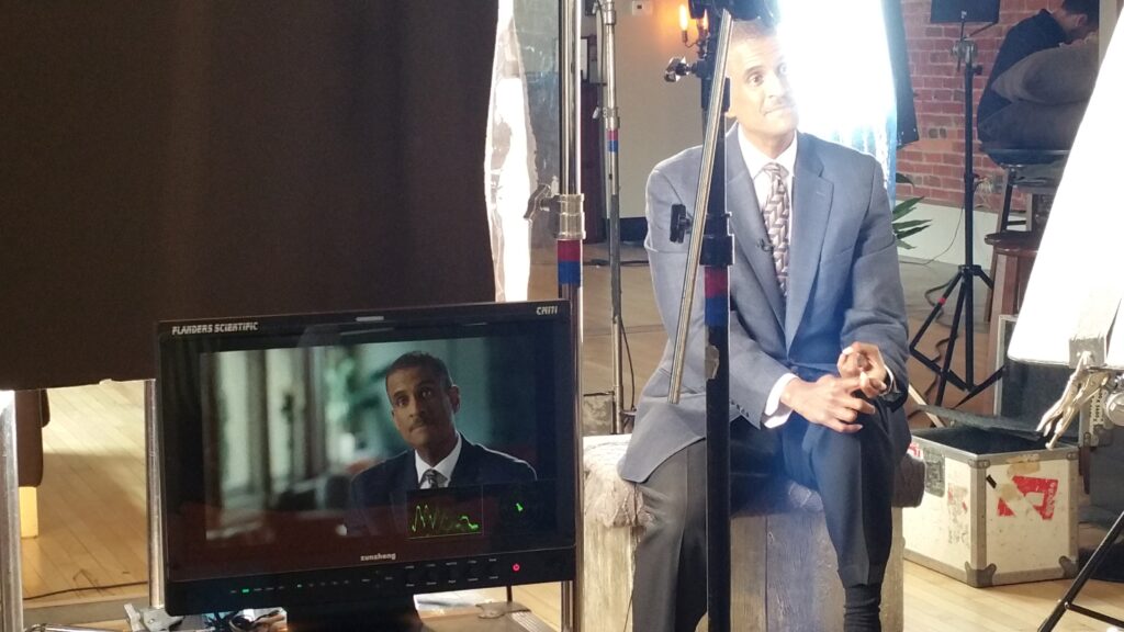 A subject interview in a studio answering questions for an interview. The foreground shows a large screen monitor showing the interviewee.